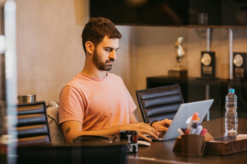 07 dicas para equilibrar a vida pessoal e a profissional Man with short hair in pink t-shirt working on a laptop in a modern office setting.