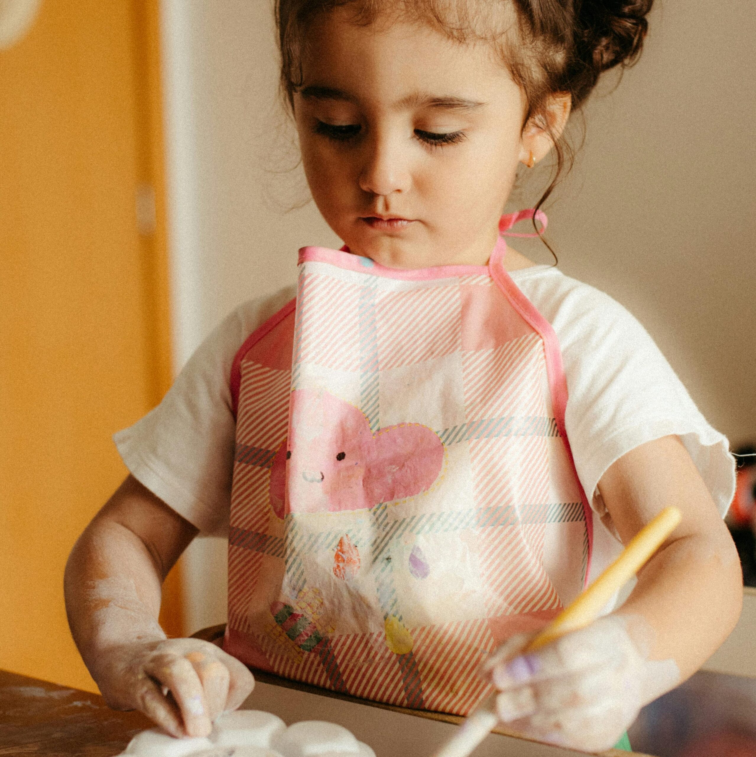 Começar o dia bem é o segredo para ter um grande dia A little girl paints a plaster mold indoors, wearing a pink apron. Ideal for art and education themes.