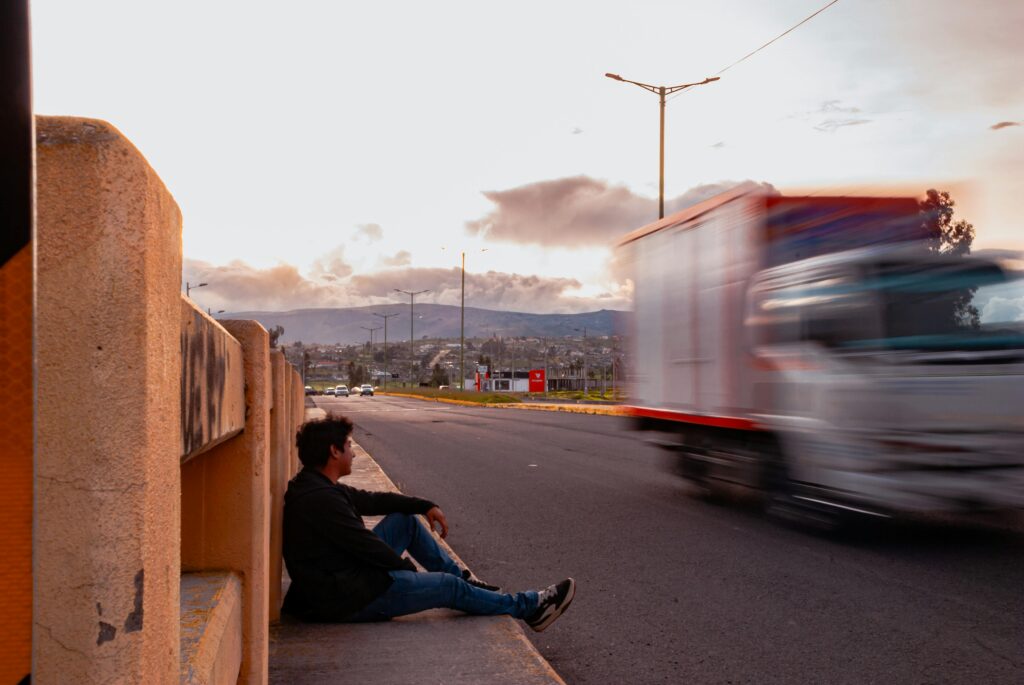 A contemplative moment as a man sits by the roadside with a truck speeding by during sunset.