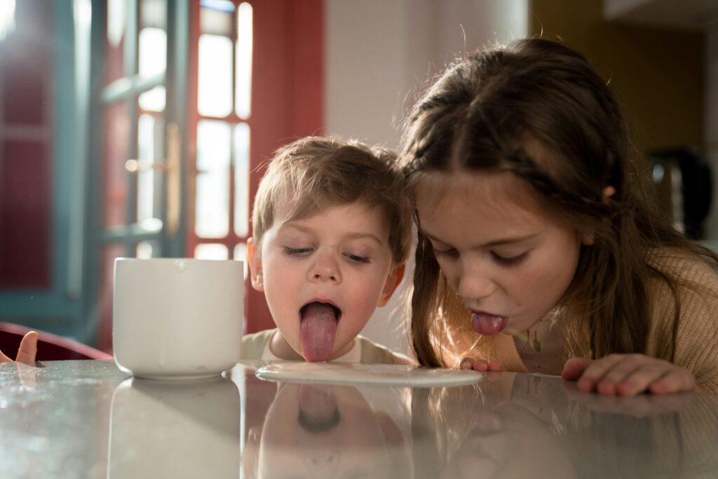 Começar o dia bem é o segredo para ter um grande dia Two adorable siblings playfully licking a table near a mug indoors.