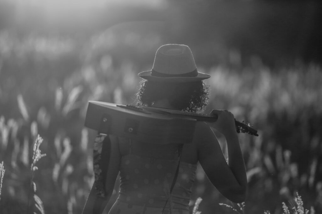 A woman walks through a sunlit field carrying a guitar, captured in black and white.