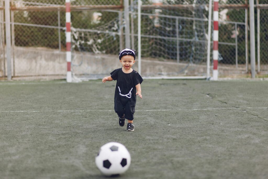 Full body cheerful laughing little boy in casual black outfit with ball walking on green empty sports ground on sunny summer day